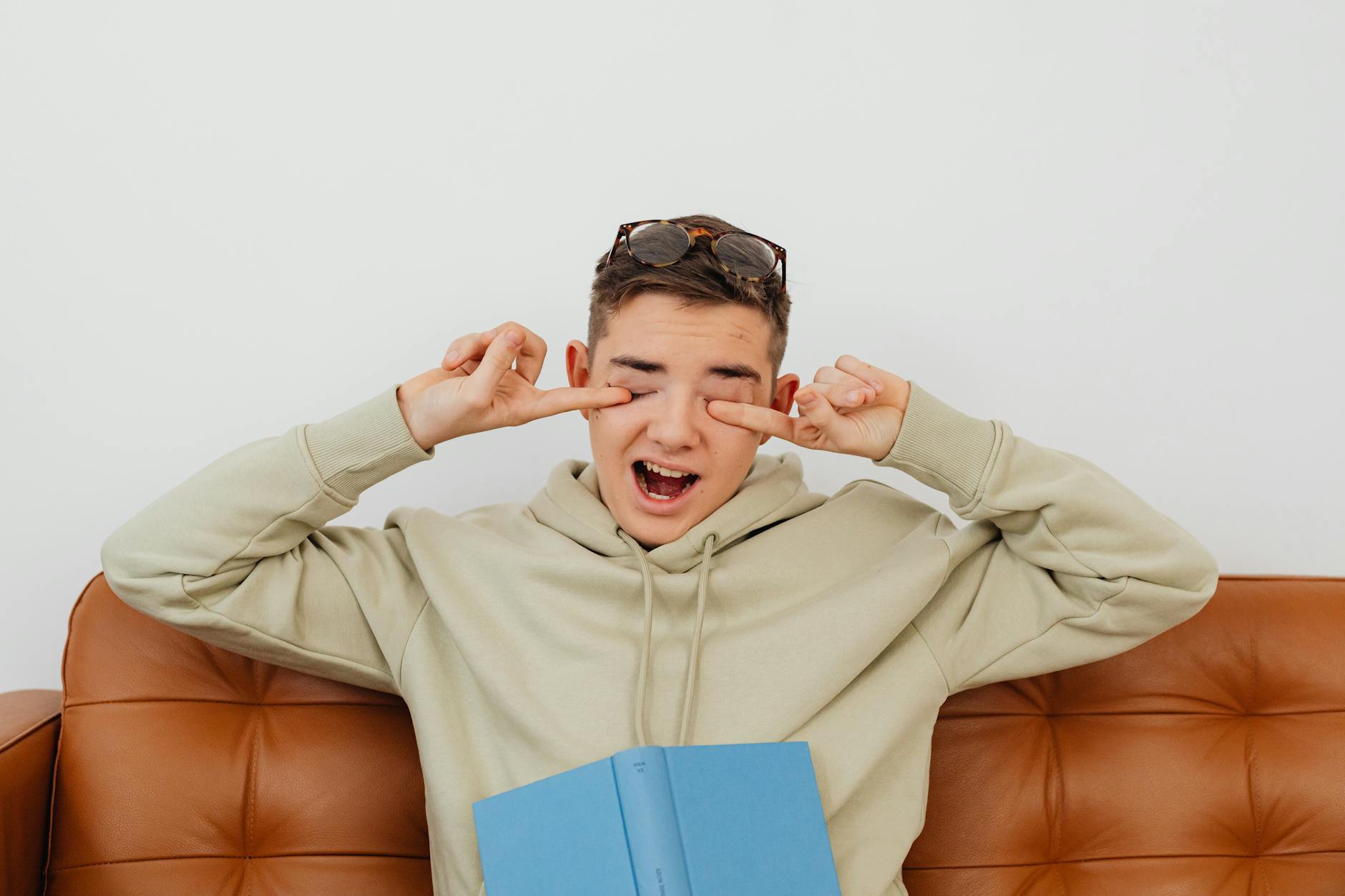 Zmęczony nastolatek w bluzie na kanapie trze suche, podrażnione oczy